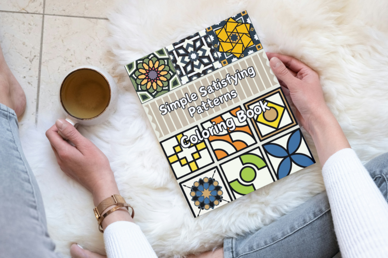 Person holding a Simple Satisfying Patterns Coloring Book while relaxing with a cup of tea on a soft rug.