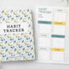 Habit tracker notebook with botanical spiral cover displayed next to a weekly habit tracking page on a minimalist desk