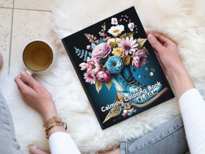 A physical copy of the Flowers Bloom Calming Coloring Book on a wooden side table next to a cactus.